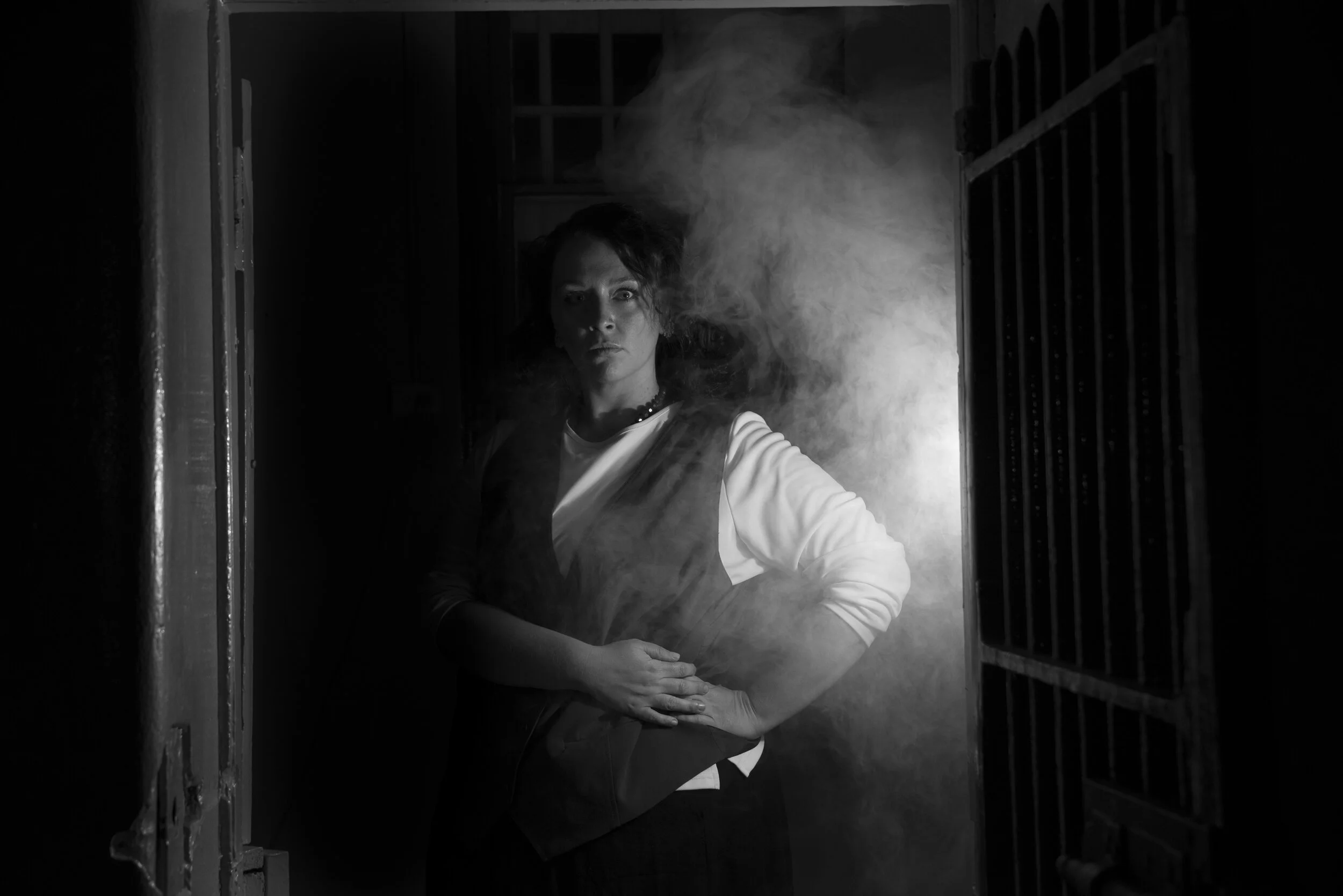 Black-and-white Assassins portrait of a performer standing in a smoky gaol doorway.