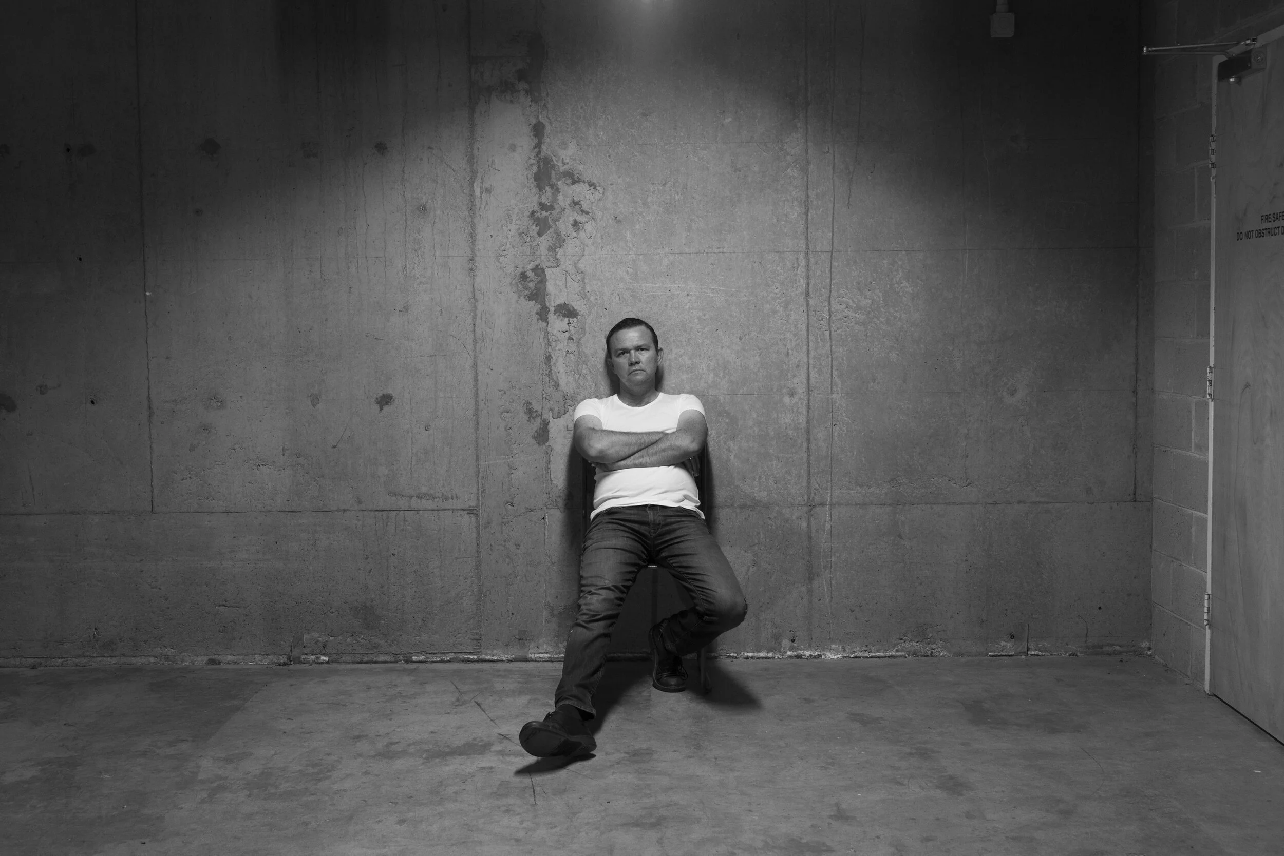 Black-and-white Assassins portrait of a performer leaning against a bare gaol cell wall.