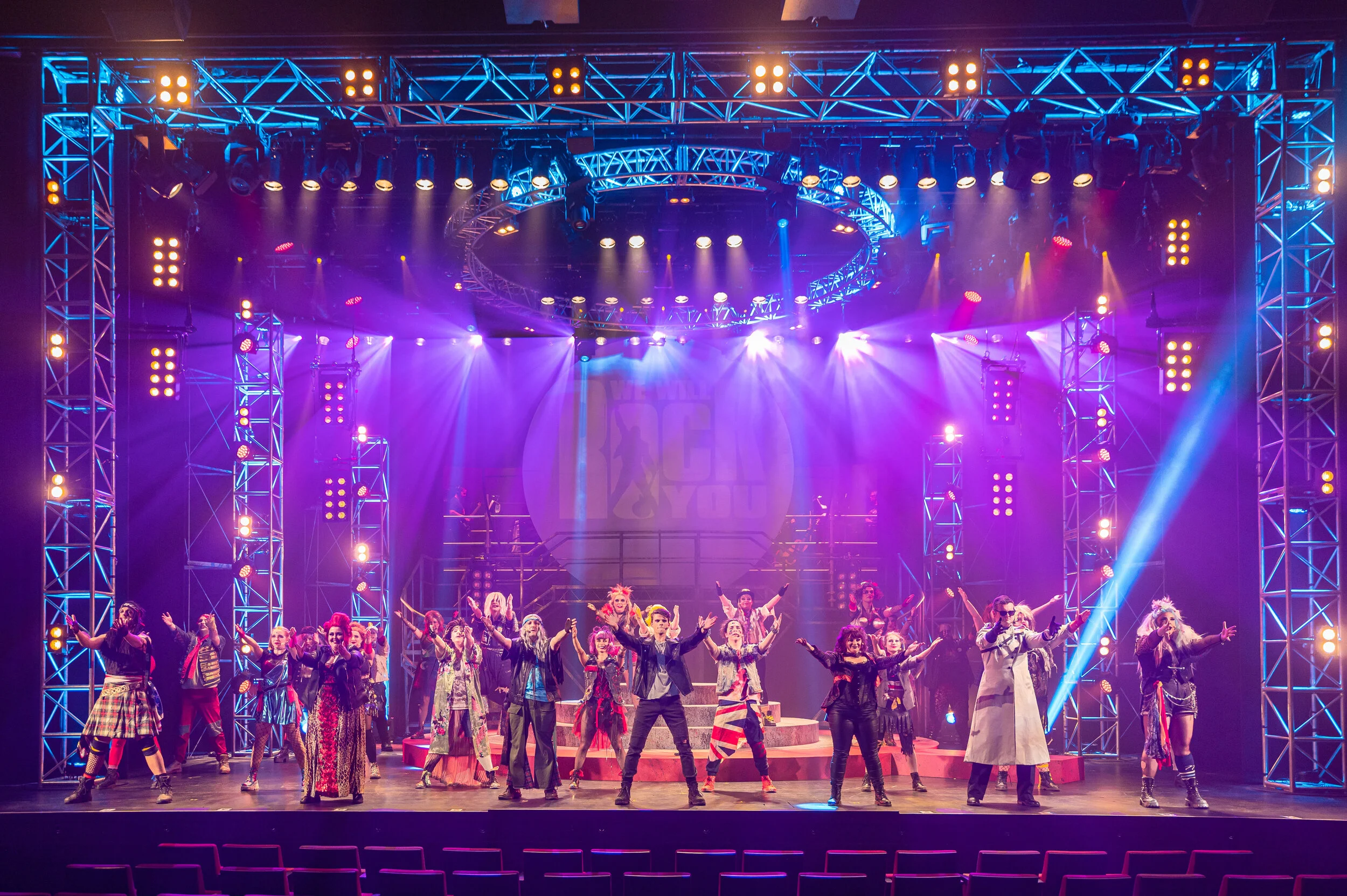 We Will Rock You ensemble beneath purple beams and a large lighting truss.