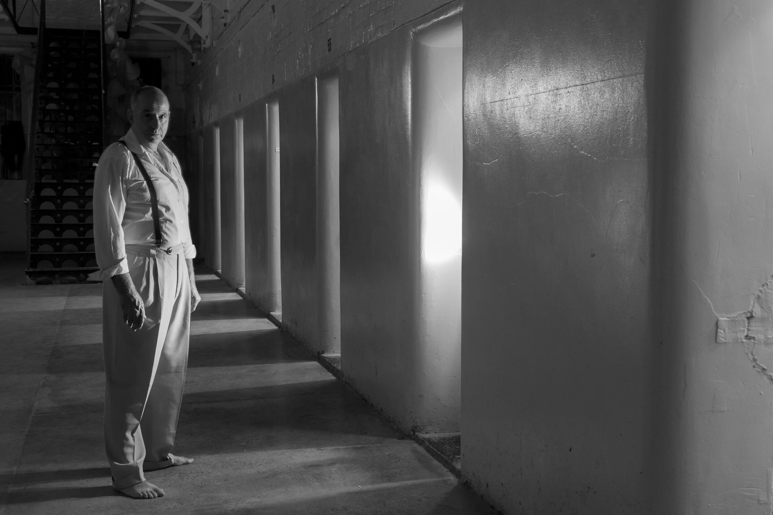 Black-and-white Assassins portrait of a performer in a gaol corridor crossed by window light.