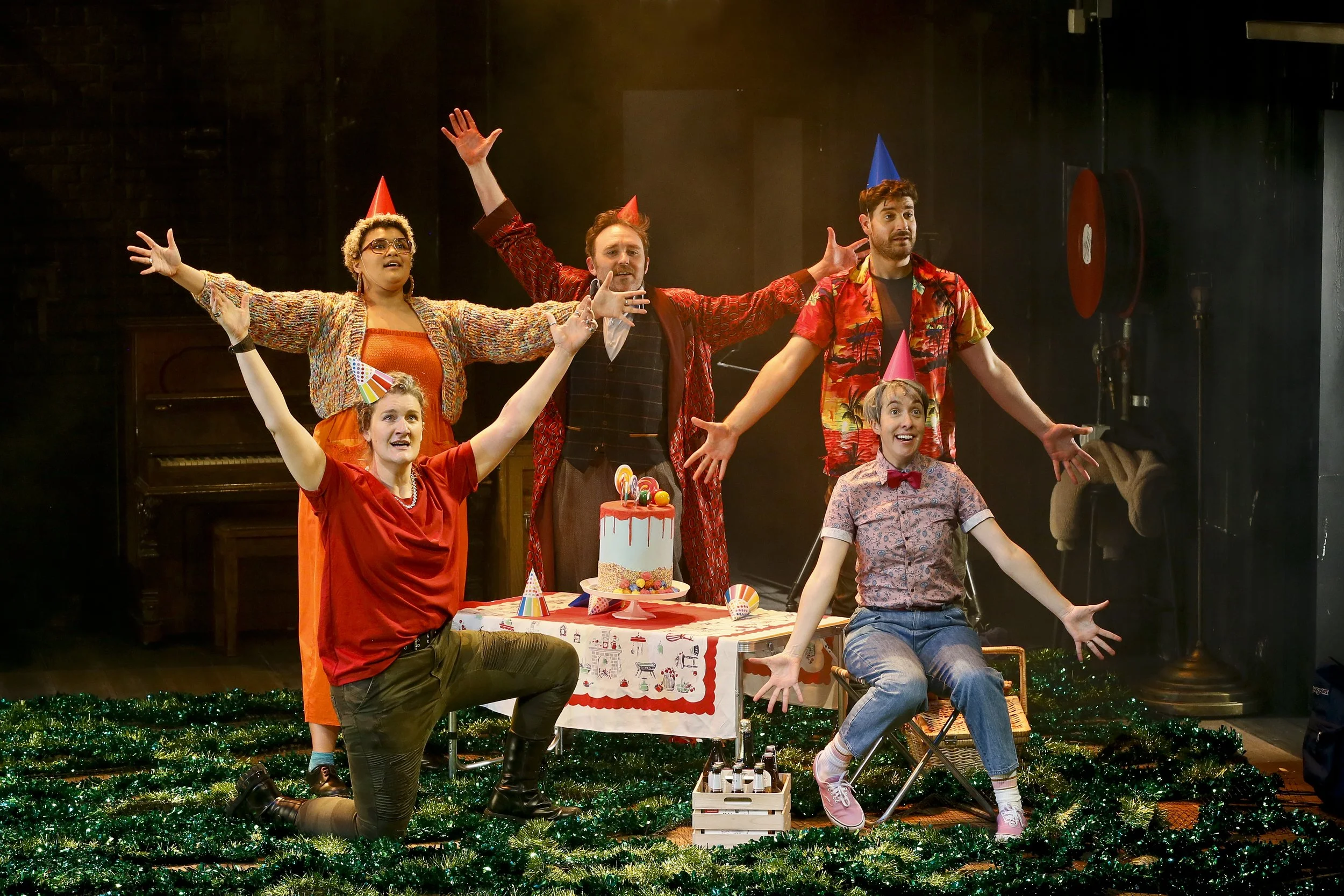 Cyrano performers wearing party hats gather around a birthday cake on a foliage-covered stage.