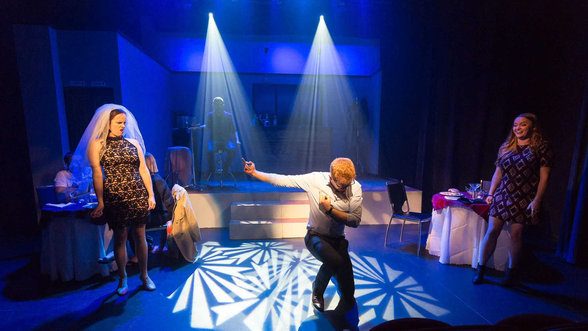 Three First Date performers on a stage lit by blue spotlights and patterned floor lighting.