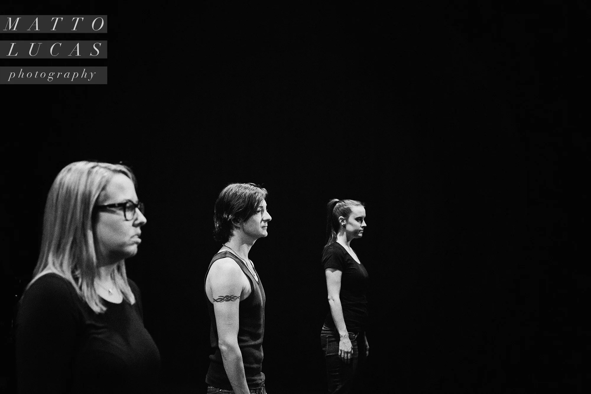 Black-and-white photograph of three [title of show] performers standing in a line on a dark stage.