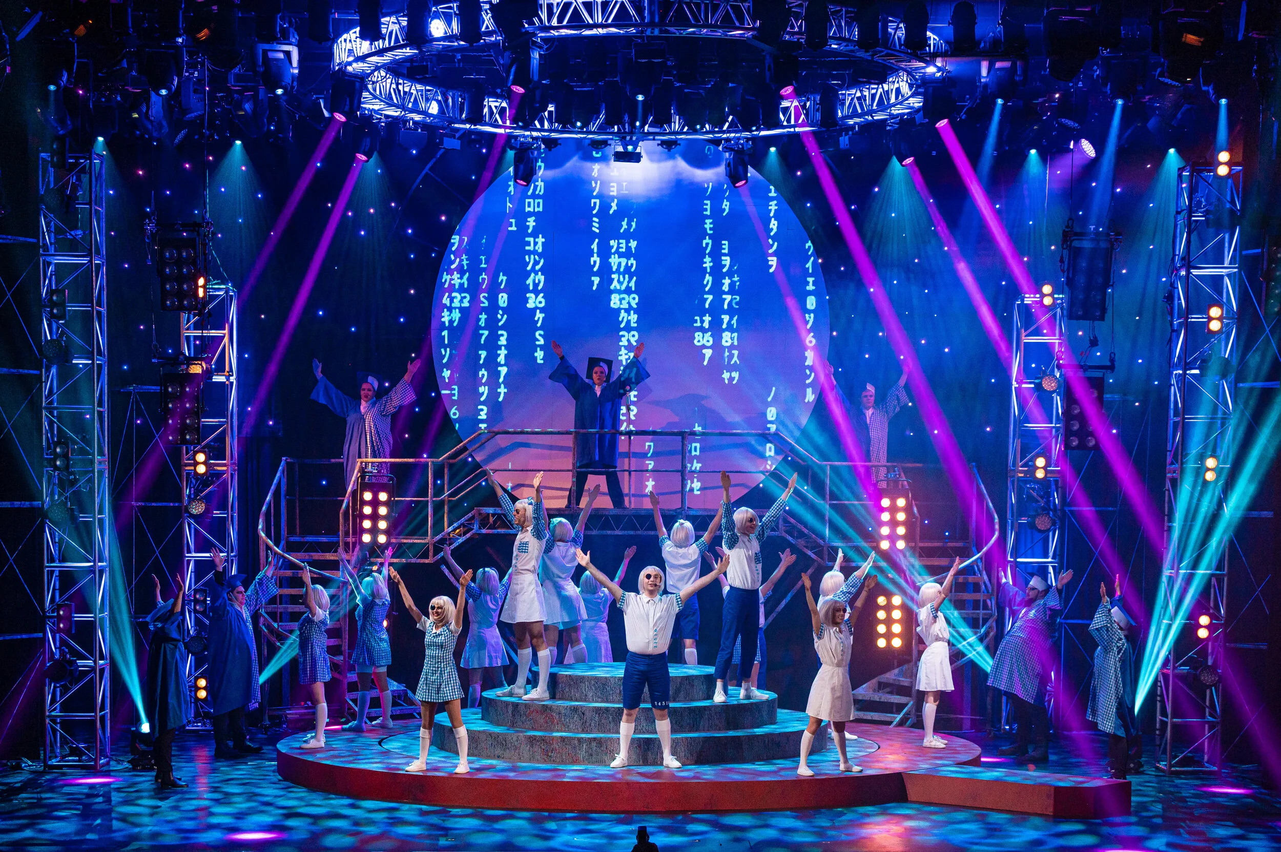 We Will Rock You performers on a raised circular platform beneath blue and magenta lighting.