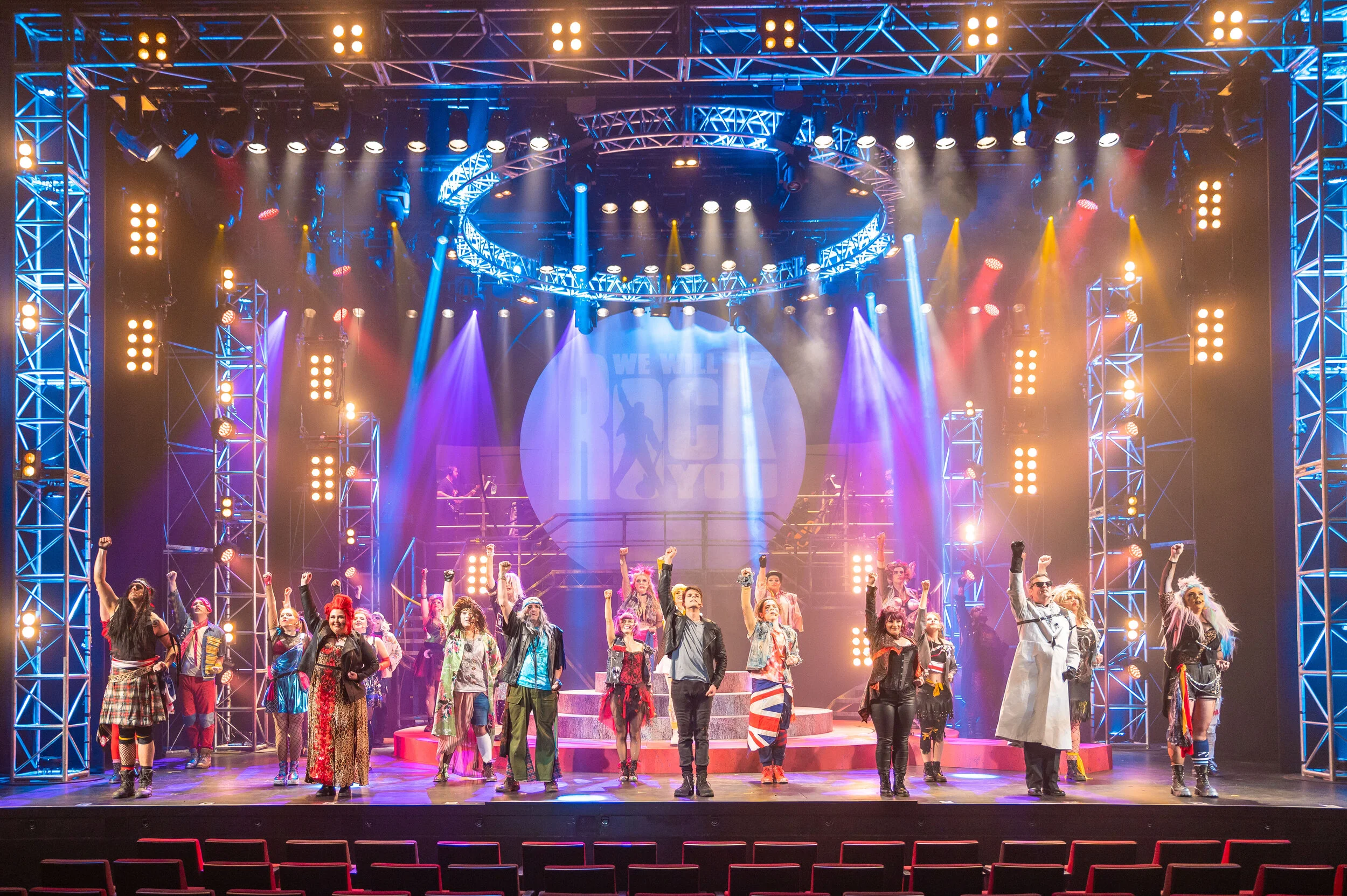 We Will Rock You ensemble across the stage beneath multicoloured lighting and a circular backdrop.