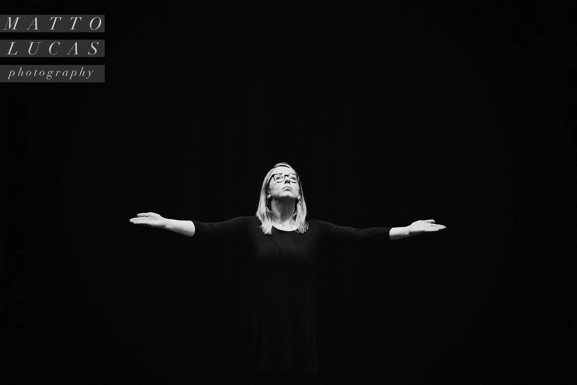 Black-and-white photograph of a [title of show] performer with arms outstretched on a dark stage.