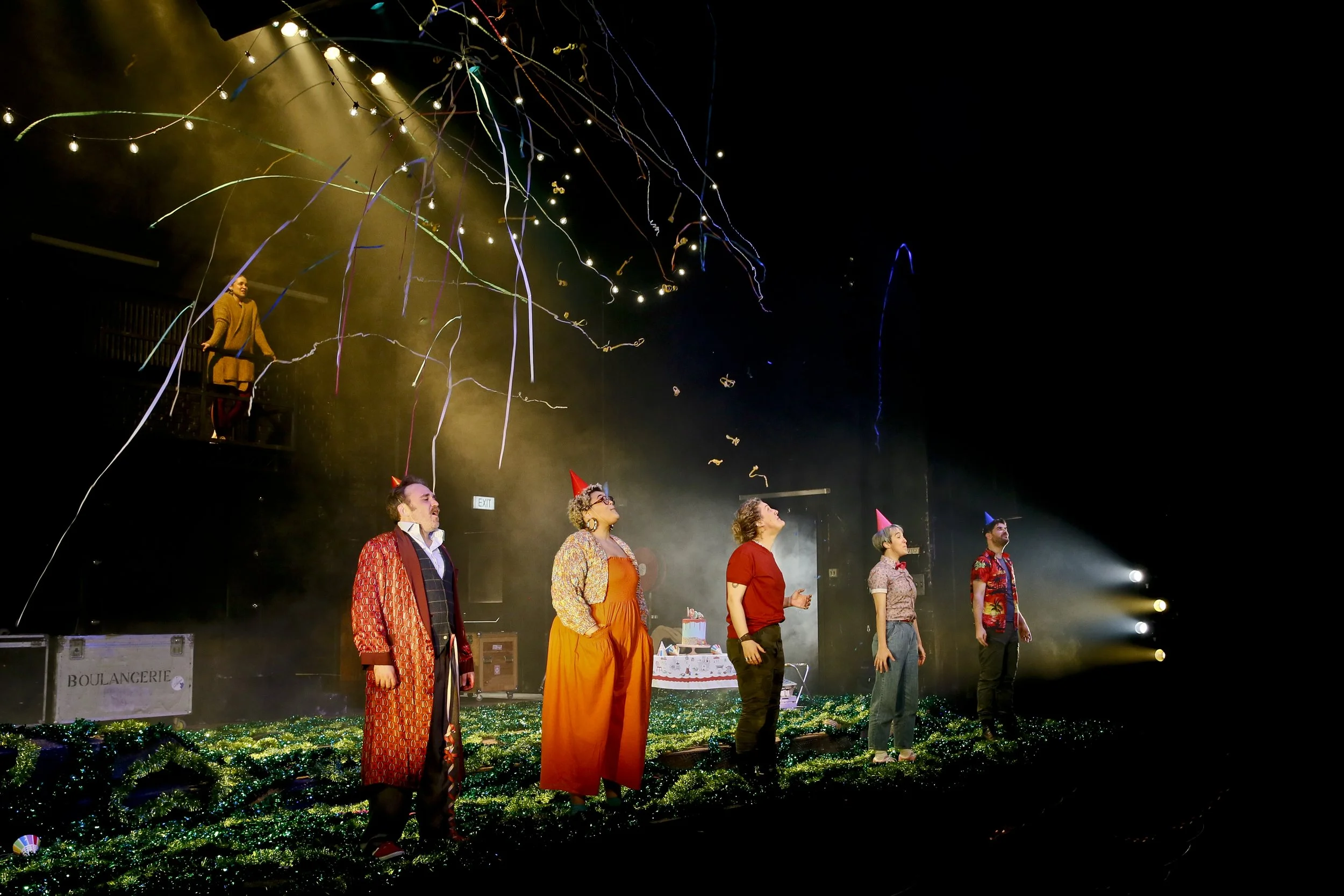 Cyrano performers beneath a suspended ring of lights on a foliage-covered stage.